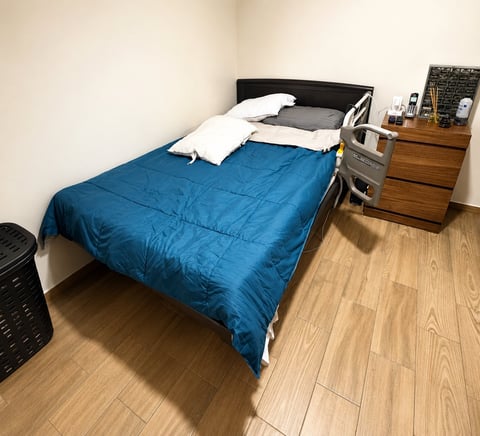 A bedroom with a bed covered in blue bedding and white pillows, wooden dresser, and hardwood flooring