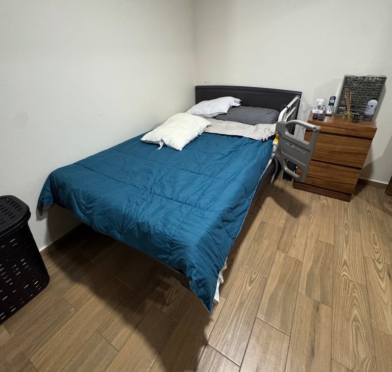 Bedroom with blue comforter on bed, white pillows, wooden nightstand, and wooden flooring in small room