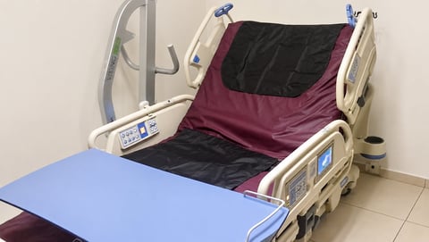 Hospital bed with burgundy and black bedding, metal side rails, and medical equipment in a clinical room with tile flooring.