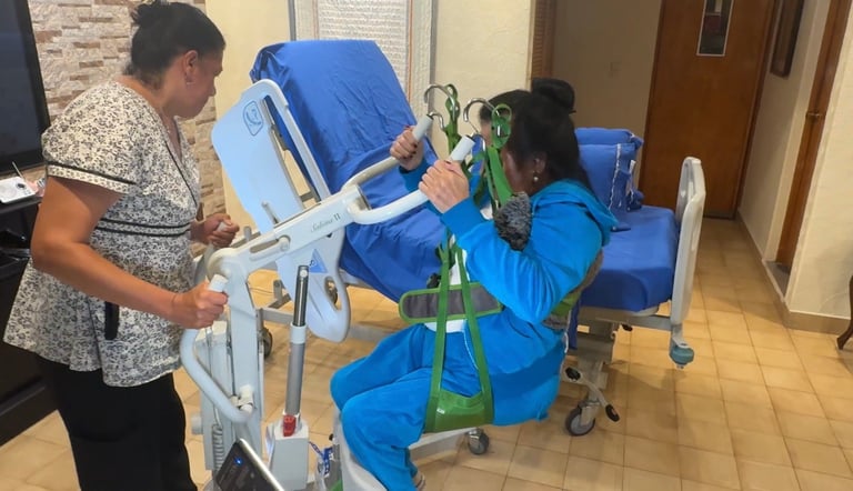 Two women assisting a patient with a mobile lifting device in a medical facility hallway